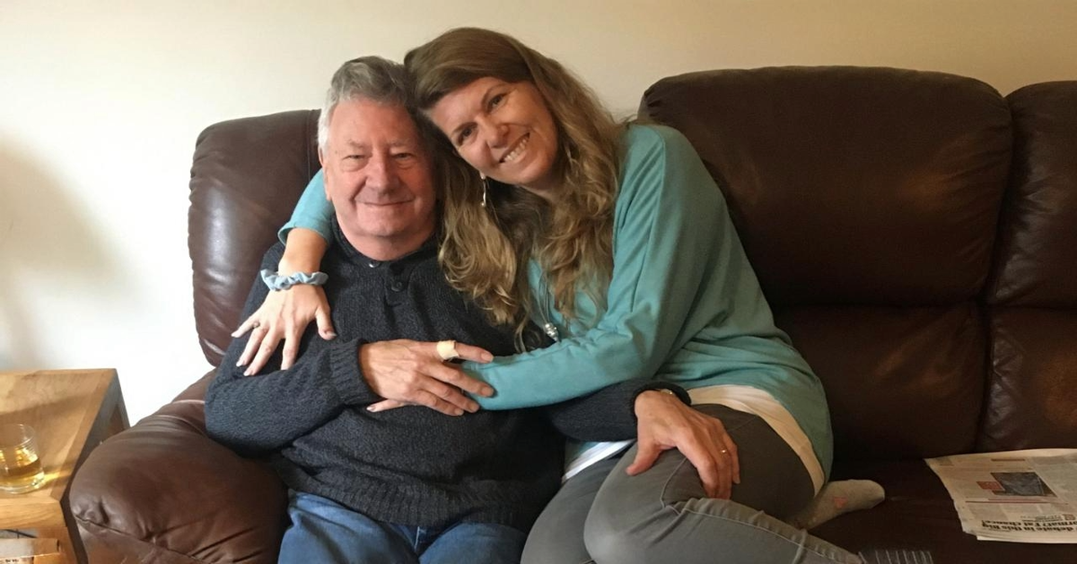 A father and daughter share a warm embrace on a leather armchair. The elderly man, dressed in a dark jumper and jeans, smiles gently, while his daughter, wearing a teal top, leans into him with affection. Their closeness suggests a bond that has softened over time, reflecting the unexpected connection that dementia has brought into their lives.