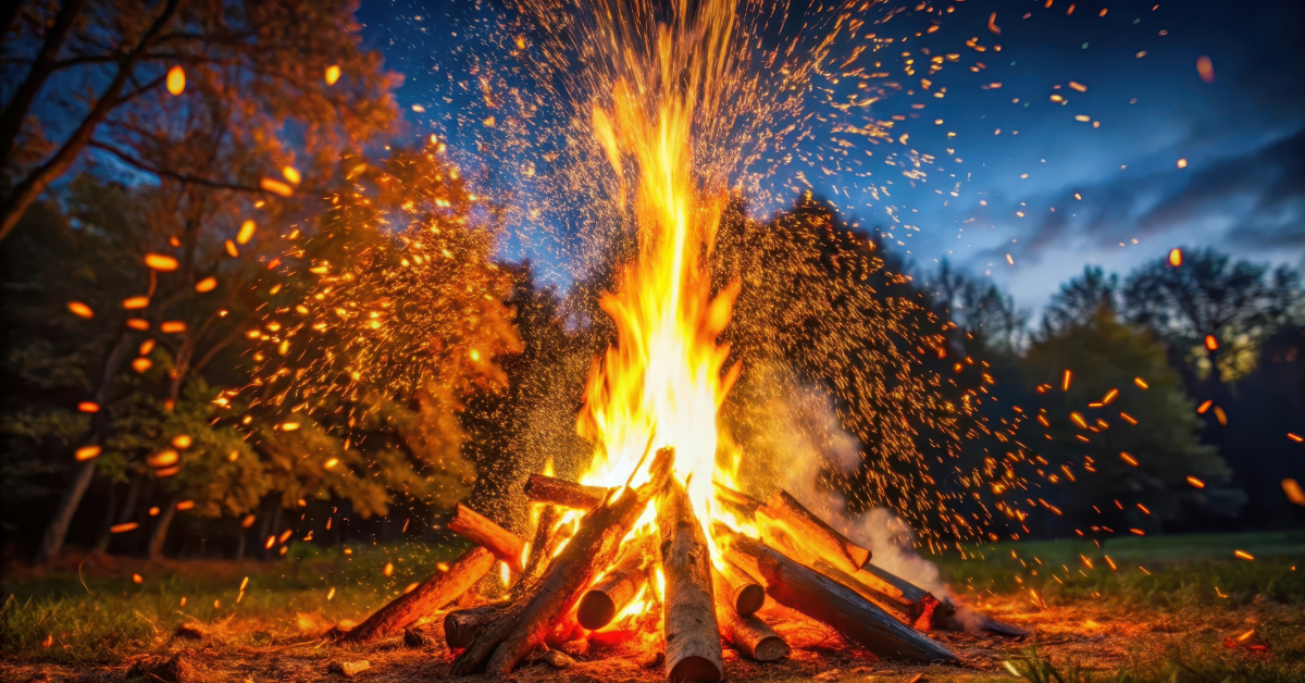 A large bonfire burning brightly at night, with glowing embers rising into the dark sky. The flames illuminate the surrounding trees, symbolising release, transformation, and the power of emotions.