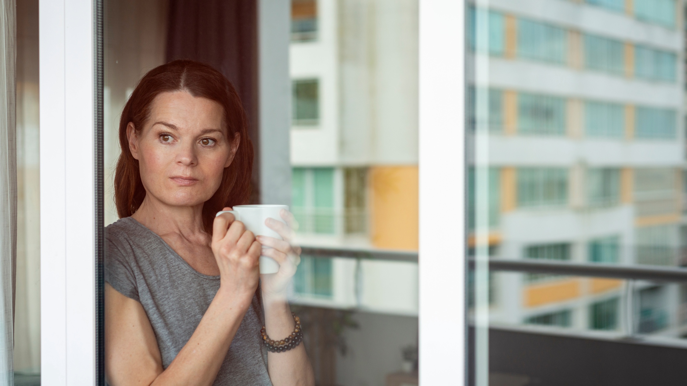 Woman standing at a window holding a mug, looking pensive and reflective, symbolising internal conflict and emotional introspection.